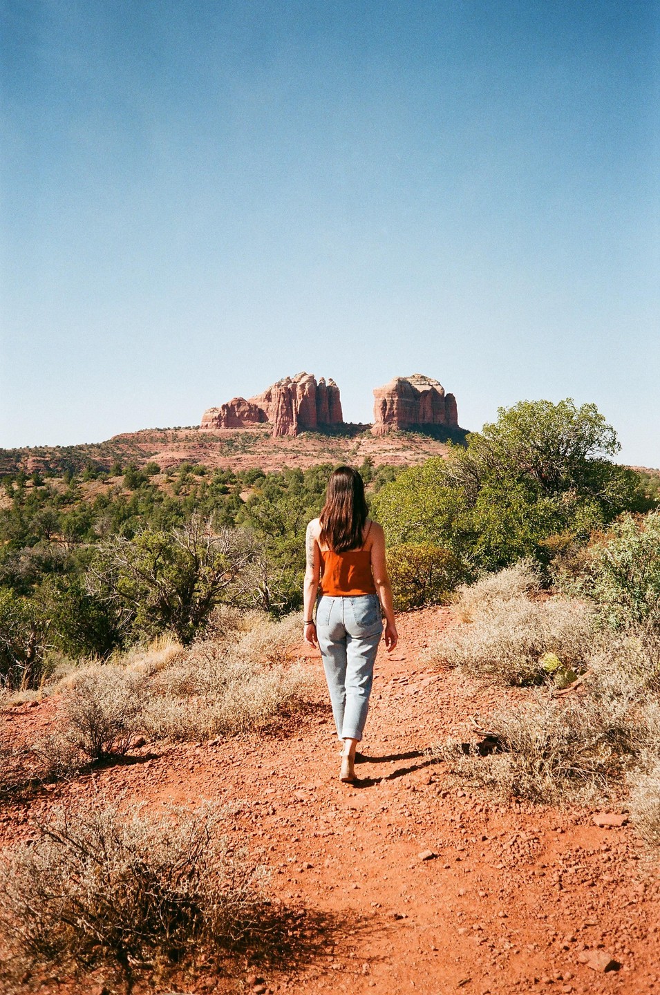 A woman walks along a red dirt path towards red rock formations under a clear blue sky.