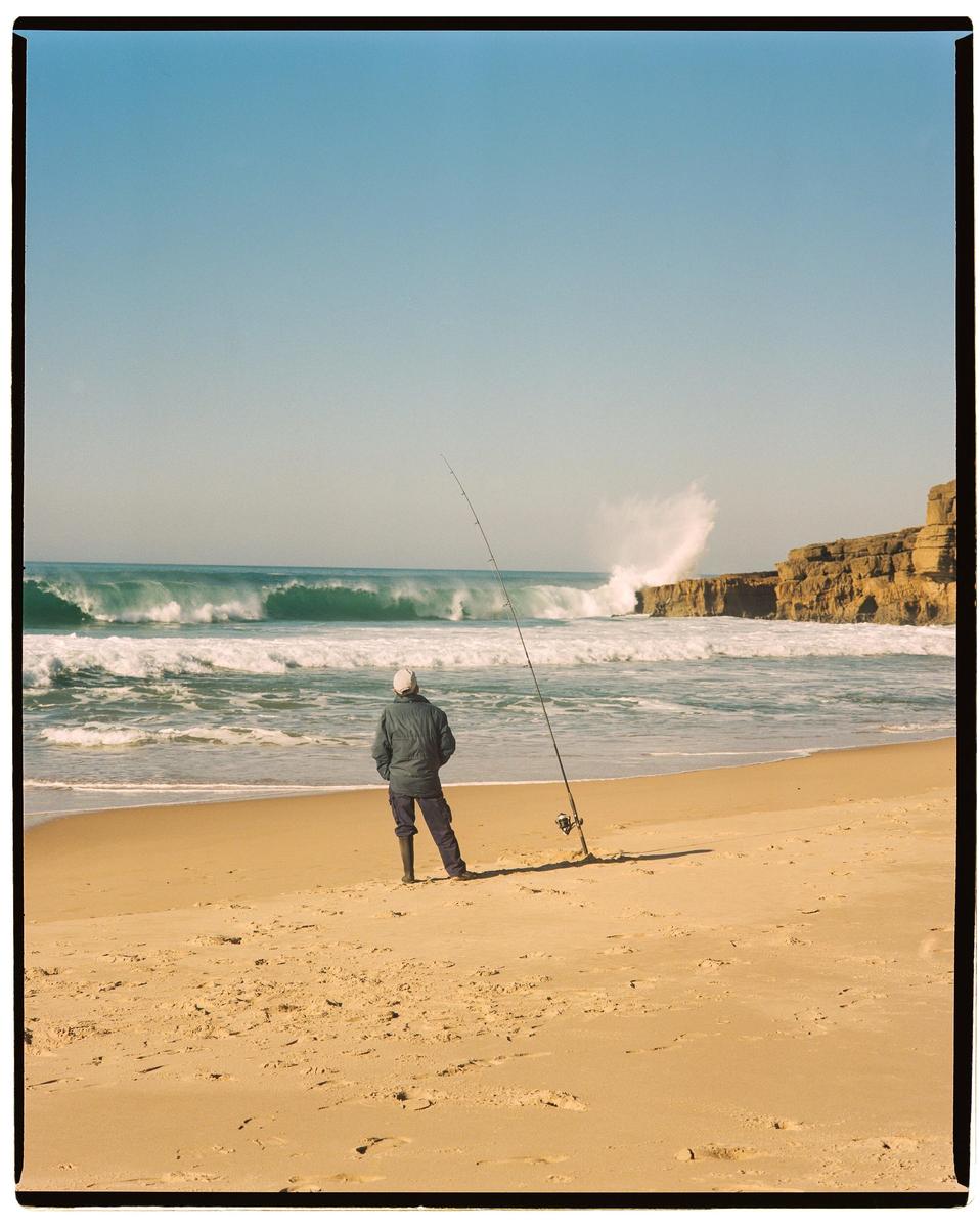 A man stands on a sandy beach with a fishing rod, looking out at large waves crashing near a rocky cliff.