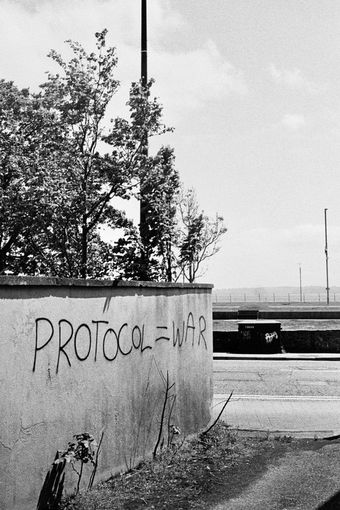 The words "PROTOCOL=WAR" are spray-painted on a concrete wall next to a road and trees.