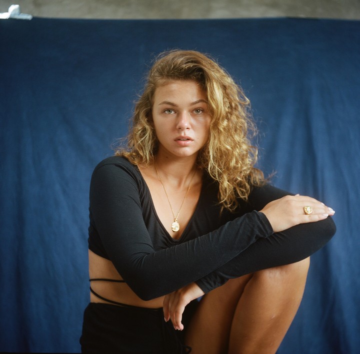 A woman with curly hair and a black top sits against a blue background.