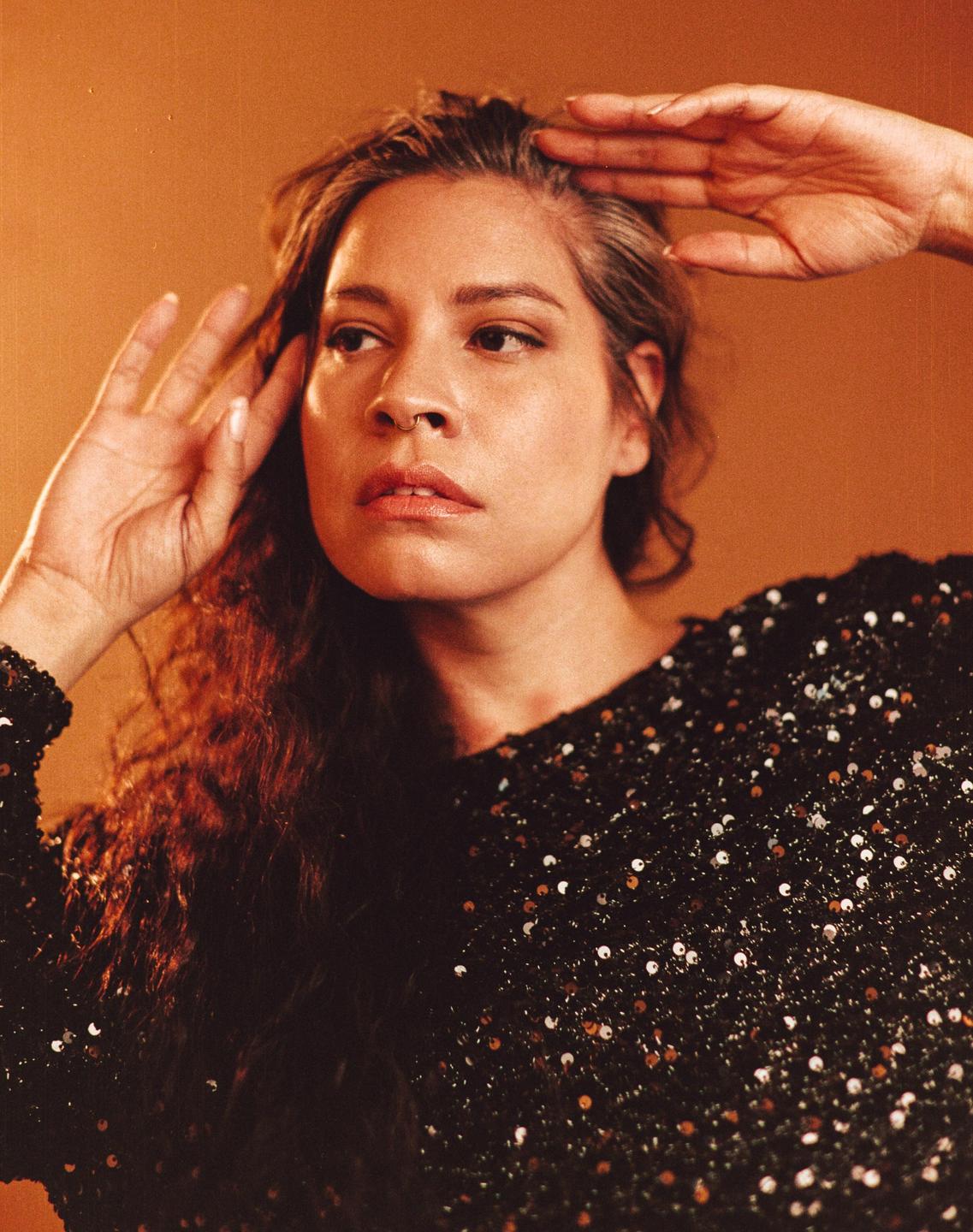 A woman with long curly hair and a sequin top poses with her hands near her head.