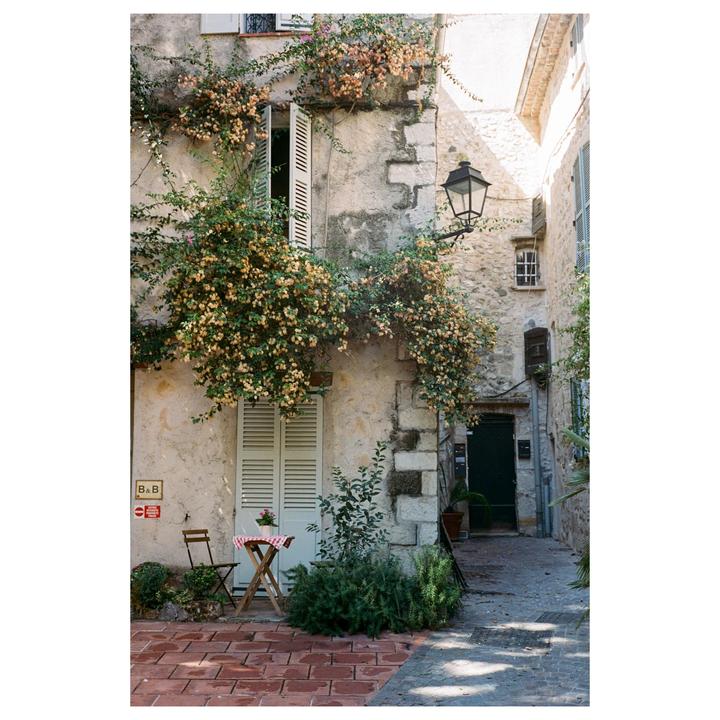 A stone building covered in flowering vines has an open window, a closed door, and a small table and chair on a brick patio.