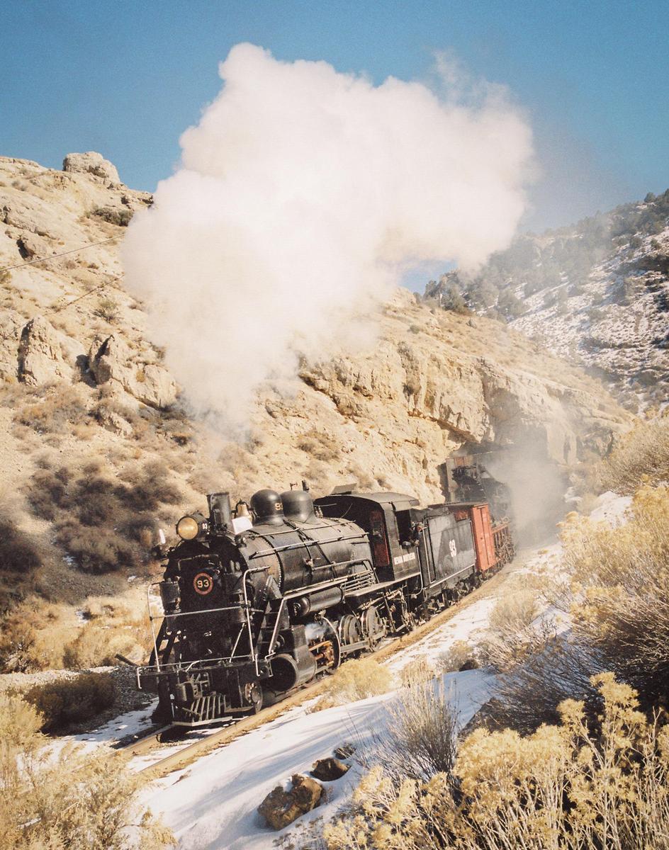 A black steam locomotive travels along a snowy track through a rocky, arid landscape under a clear blue sky.