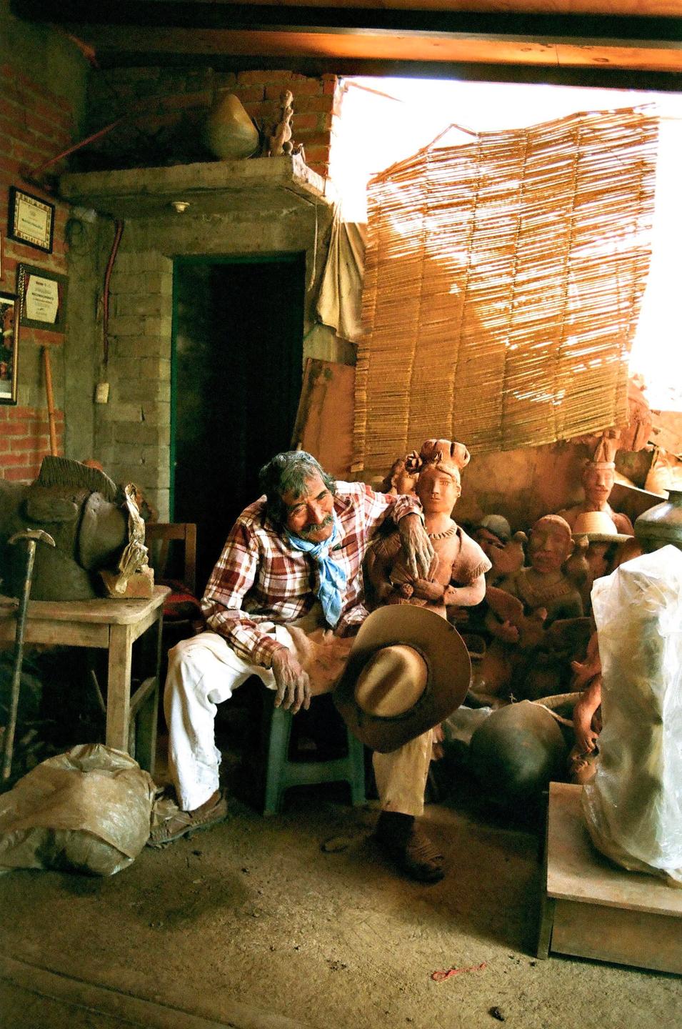 A man sits in a room filled with pottery, holding a cowboy hat.