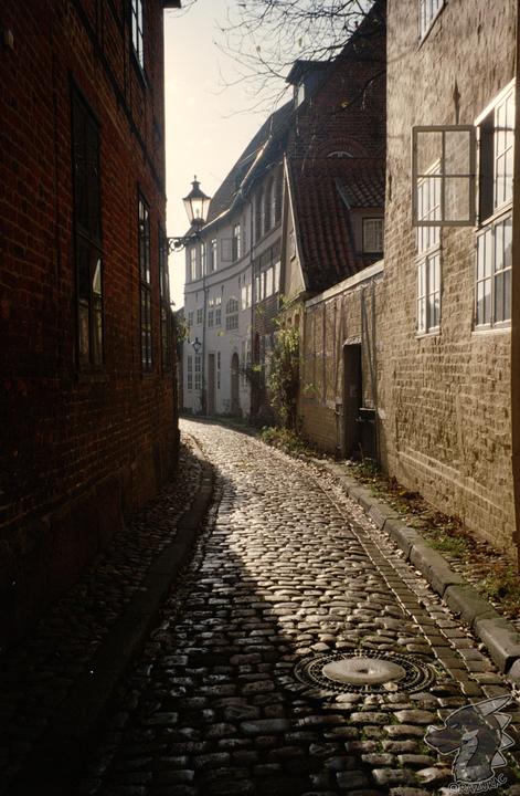 A narrow cobblestone street curves between brick buildings with a street lamp visible in the distance.