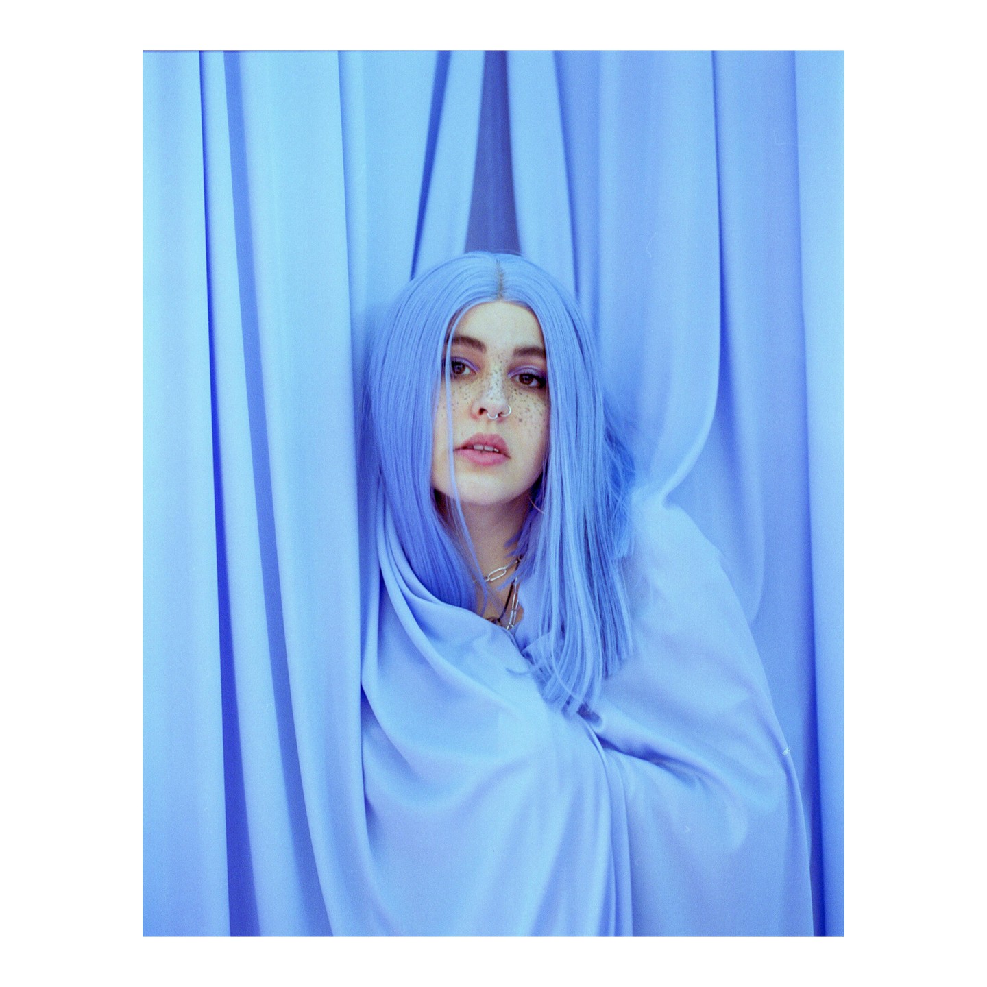 A woman with blue hair and freckles is wrapped in blue fabric, peeking from behind blue curtains.