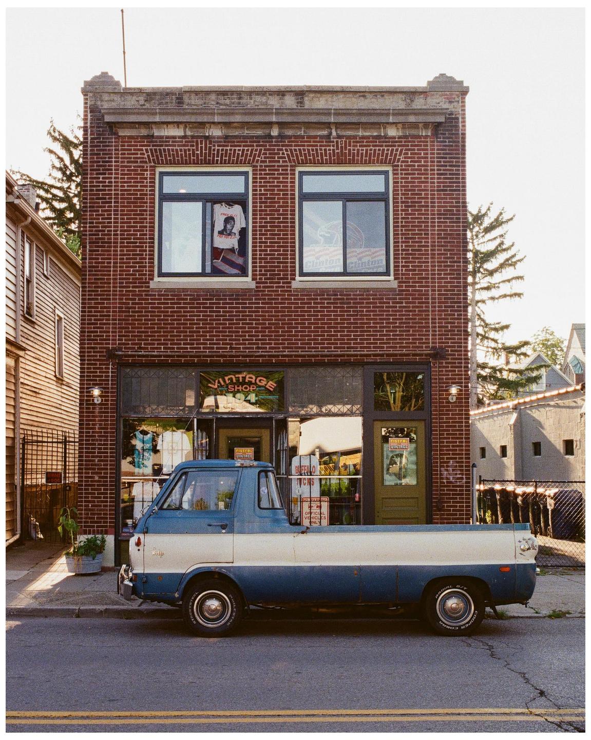 A blue and white vintage pickup truck is parked on a street in front of a brick building with a vintage shop.