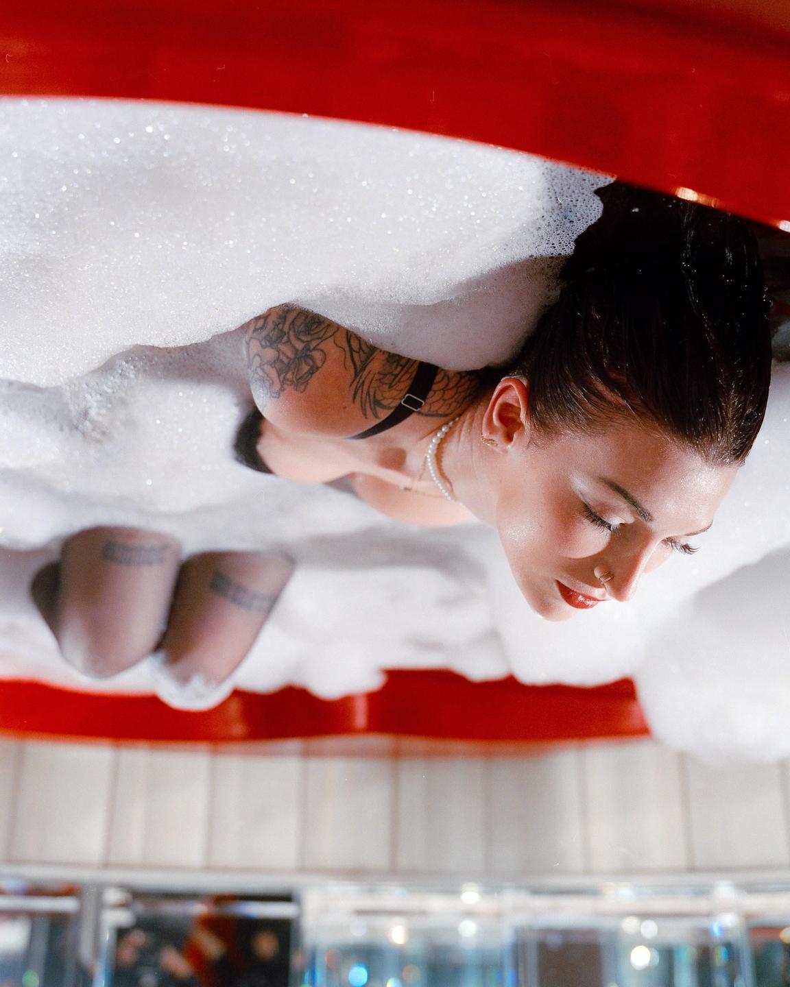 A woman with red lipstick and a pearl necklace is submerged in a bubble bath, with her tattooed arm visible.