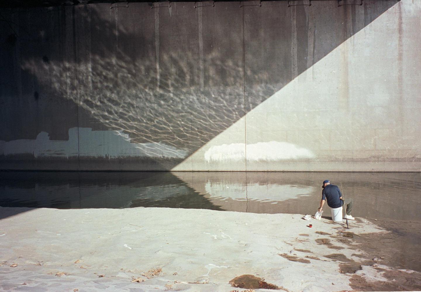 A man is fishing in a concrete river with light reflecting off the water onto the wall.