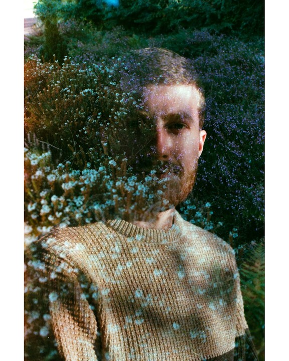 A man with a beard is visible through a double exposure with flowering bushes.