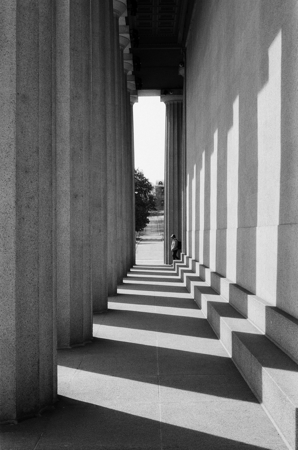 A man stands in the shadow of a colonnade with a street and trees visible in the distance.
