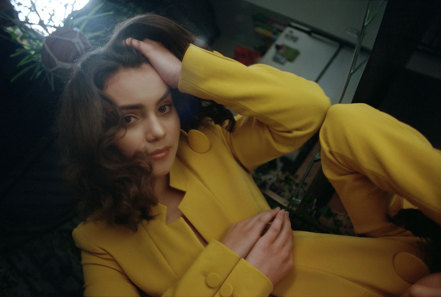 A woman in a yellow suit poses with her hand on her head, reflected in a mirror.