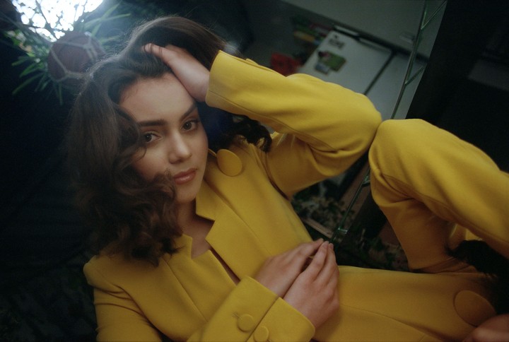 A woman in a yellow suit poses with her hand on her head, reflected in a mirror.