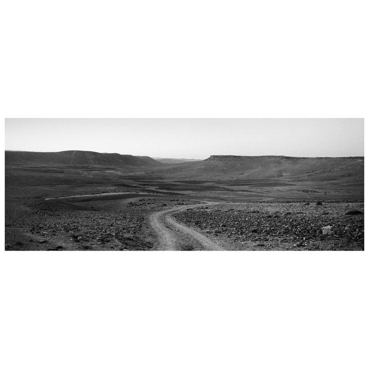 A winding dirt road leads through a rocky, arid landscape with hills in the distance.