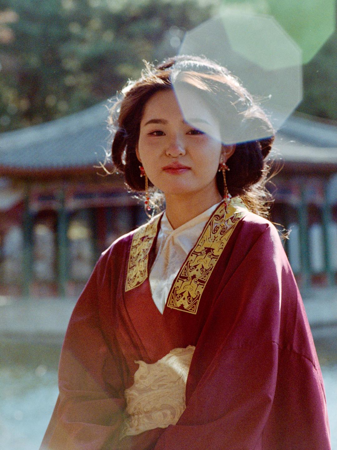A woman in traditional attire stands outdoors with a building and water in the background.