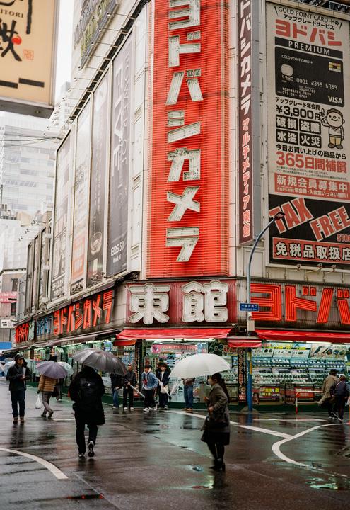 A street in Tokyo is filled with people holding umbrellas, walking past buildings with large signs on a rainy day.