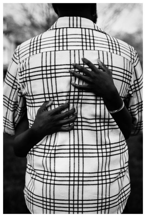 A person wearing a plaid shirt is embraced from behind by another person whose hands are visible.
