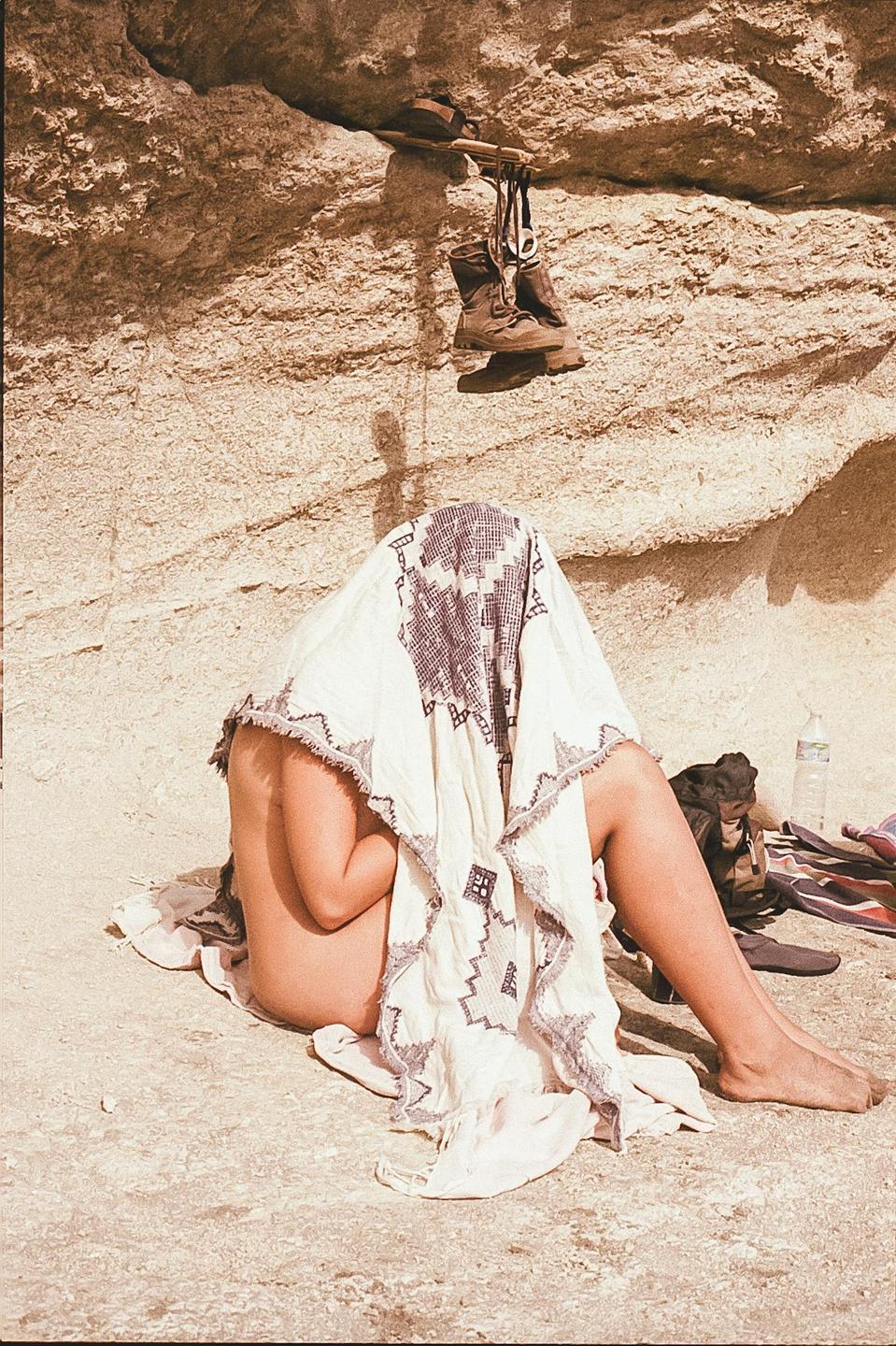 A person sits on the ground with a patterned blanket over their head, next to a rock wall with boots hanging from it.
