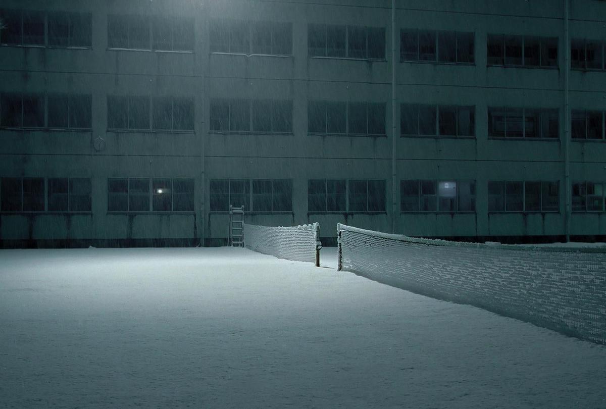 A snow-covered tennis court and building are illuminated by a single light at night.