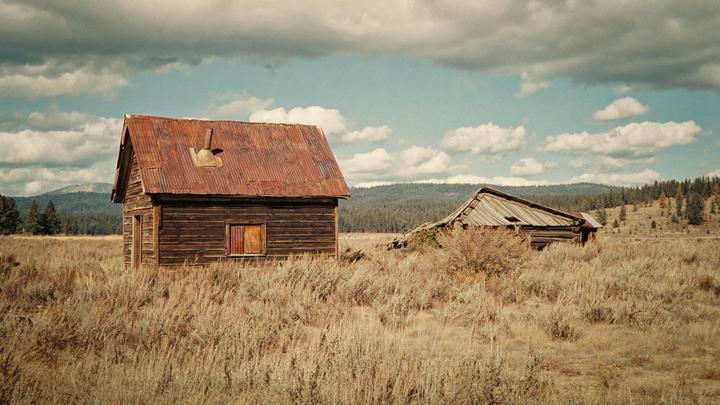 Two abandoned wooden cabins with rusted tin roofs stand in a field of dry grass under a partly cloudy sky.