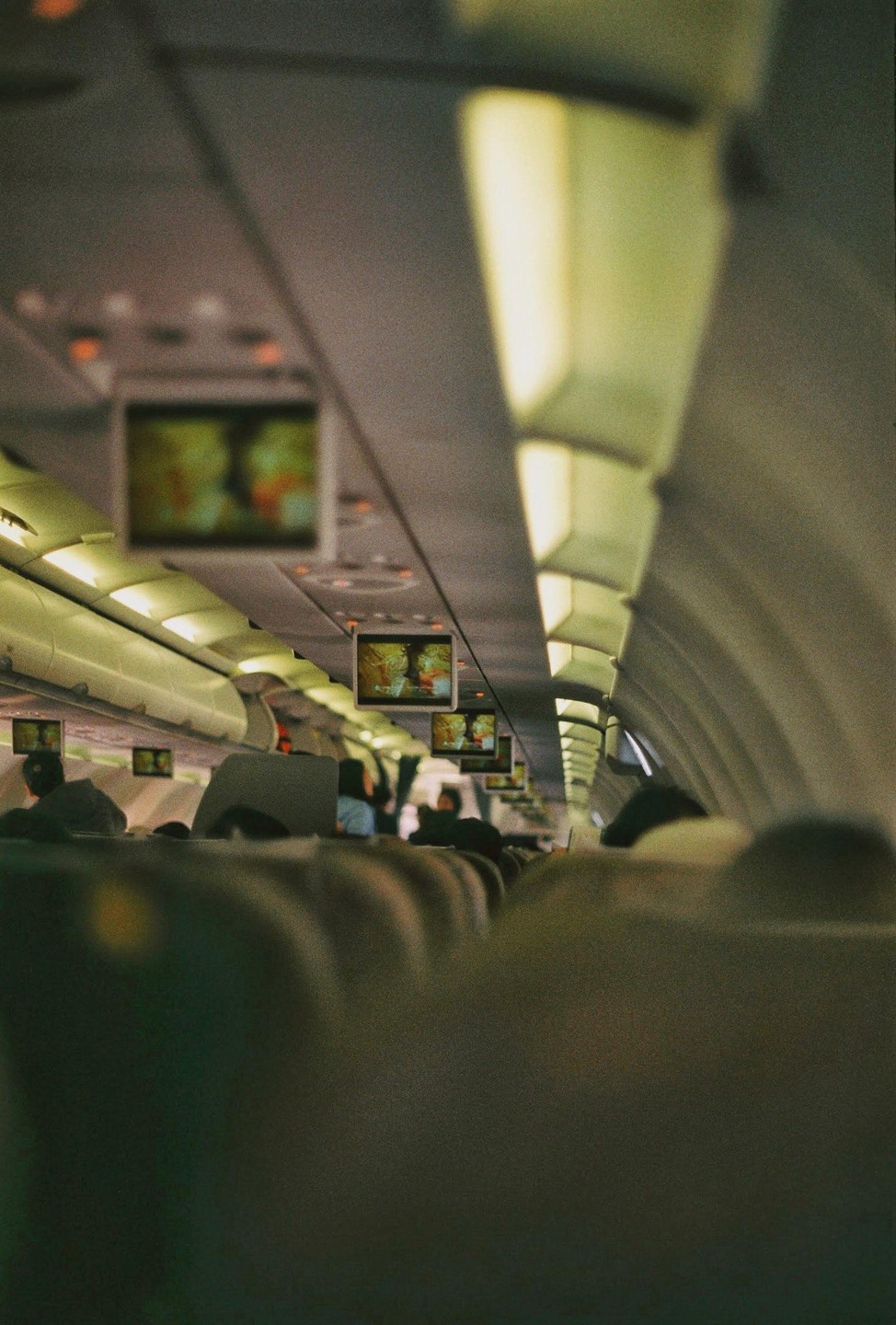 The interior of an airplane cabin is visible from a passenger's perspective, with screens displaying content above the seats.
