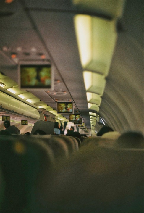 The interior of an airplane cabin is visible from a passenger's perspective, with screens displaying content above the seats.