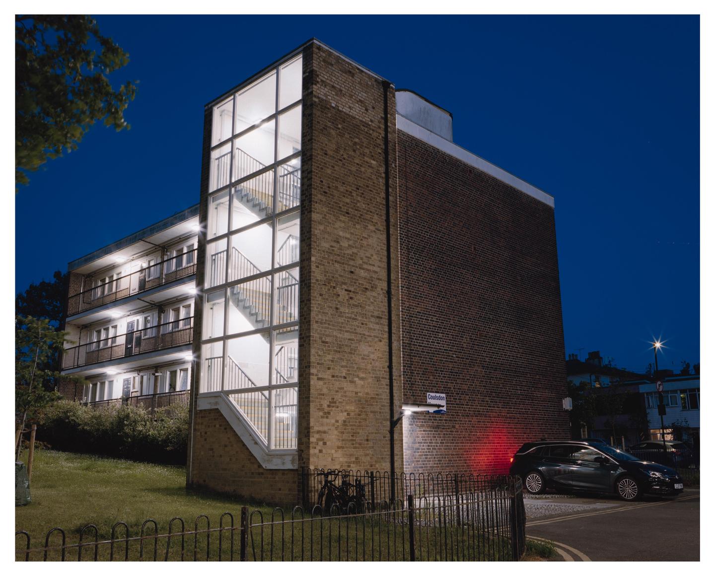A brick apartment building with an illuminated glass stairwell stands next to a street with a parked car at night.