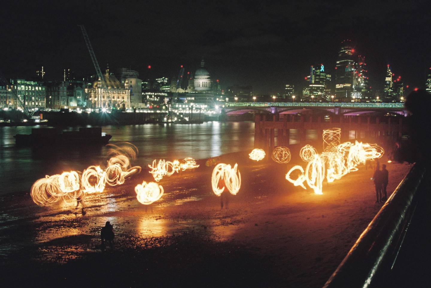 People create light trails with fire on a riverbank at night, with a city skyline and bridge in the background.