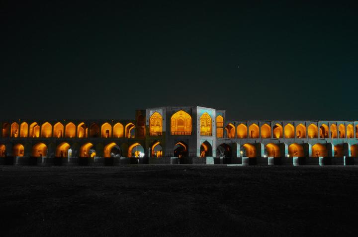 A long, ornate bridge with many arches is illuminated at night, with a dark sky above and dark ground below.