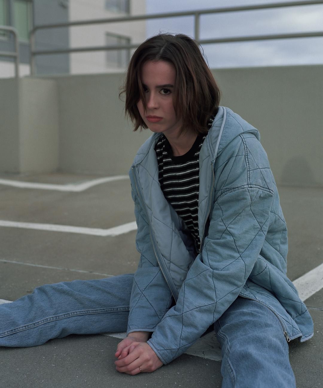 A woman with short hair sits on the ground with her legs spread apart, wearing a quilted jacket and jeans.