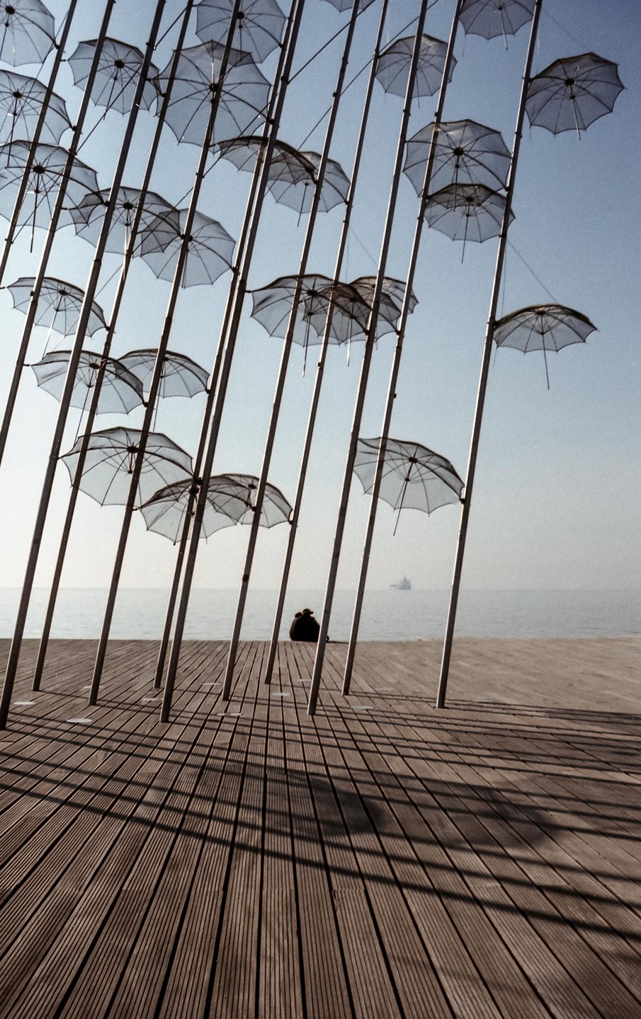 A couple sits on a wooden pier under an art installation of umbrellas, looking out at the sea.