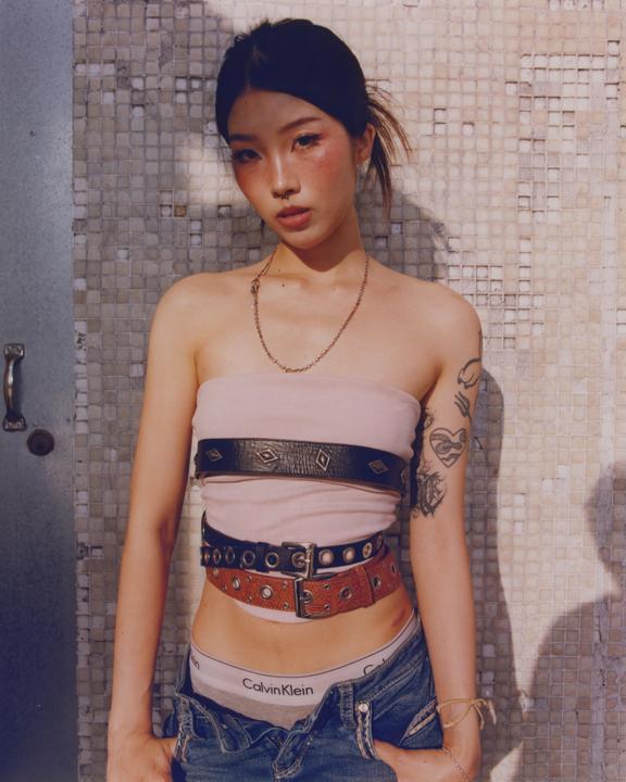 A woman with multiple belts over a pink top and jeans stands against a tiled wall.