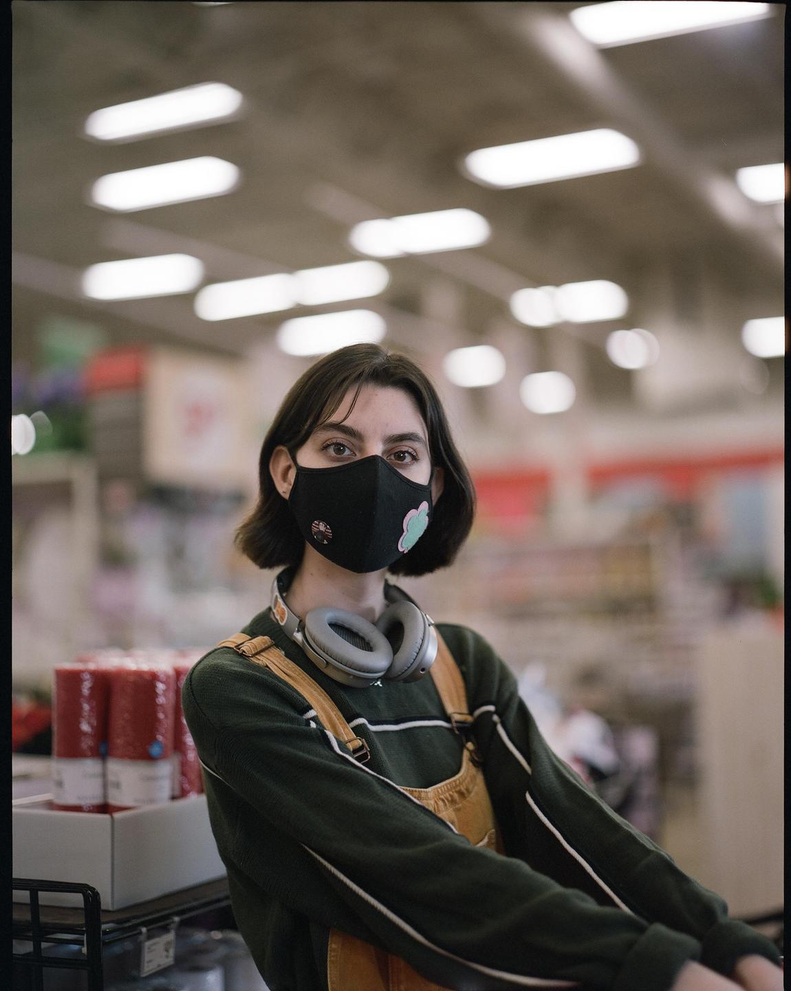 A woman wearing a face mask and headphones looks directly at the viewer in a craft store.