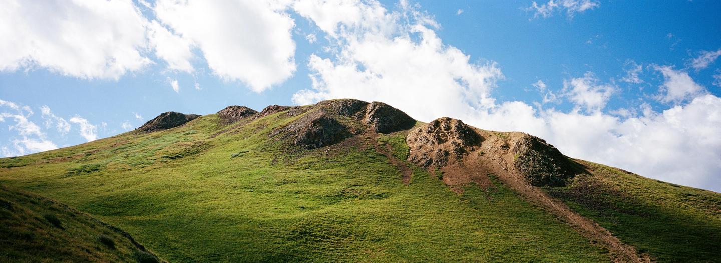 A green, grassy mountain slope with rocky outcrops is visible under a blue sky with white clouds.