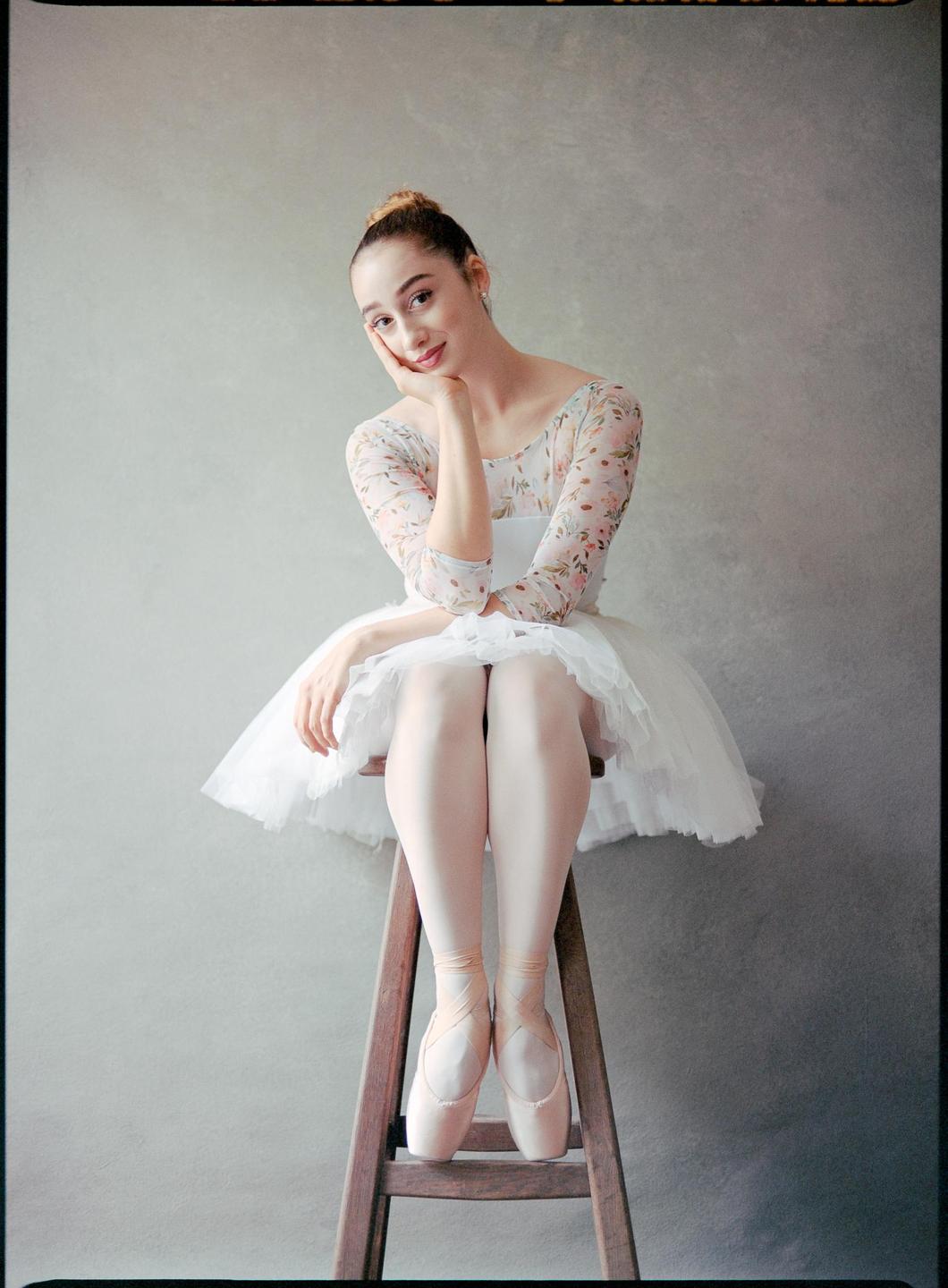 A ballerina in a white tutu and floral leotard sits on a wooden stool, resting her chin on her hand.