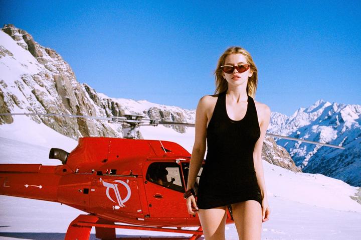 A woman in a black dress and sunglasses stands in front of a red helicopter on a snowy mountain with a clear blue sky.