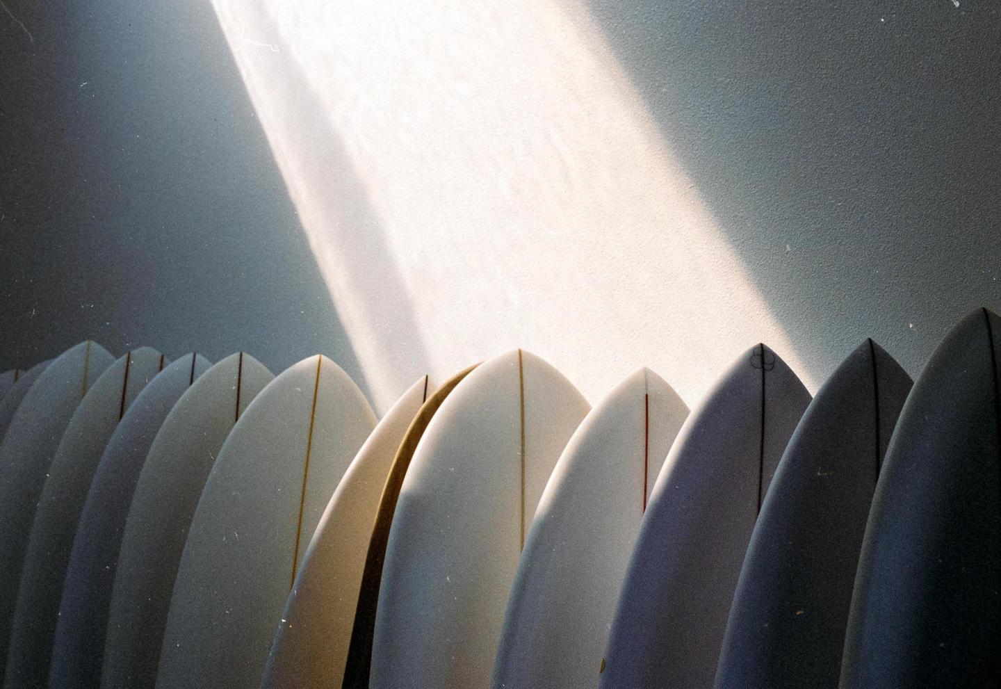 A row of surfboards stands upright against a wall, with a beam of light illuminating the upper part of the wall.