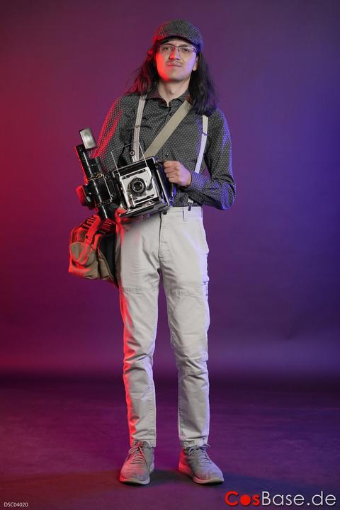 A man wearing a flat cap and suspenders holds a large format camera and a camera bag.