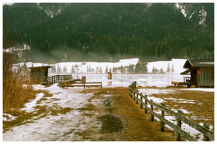 A wooden fence leads to a partially frozen lake with two small wooden structures on its banks, surrounded by snow-covered ground and a forest.