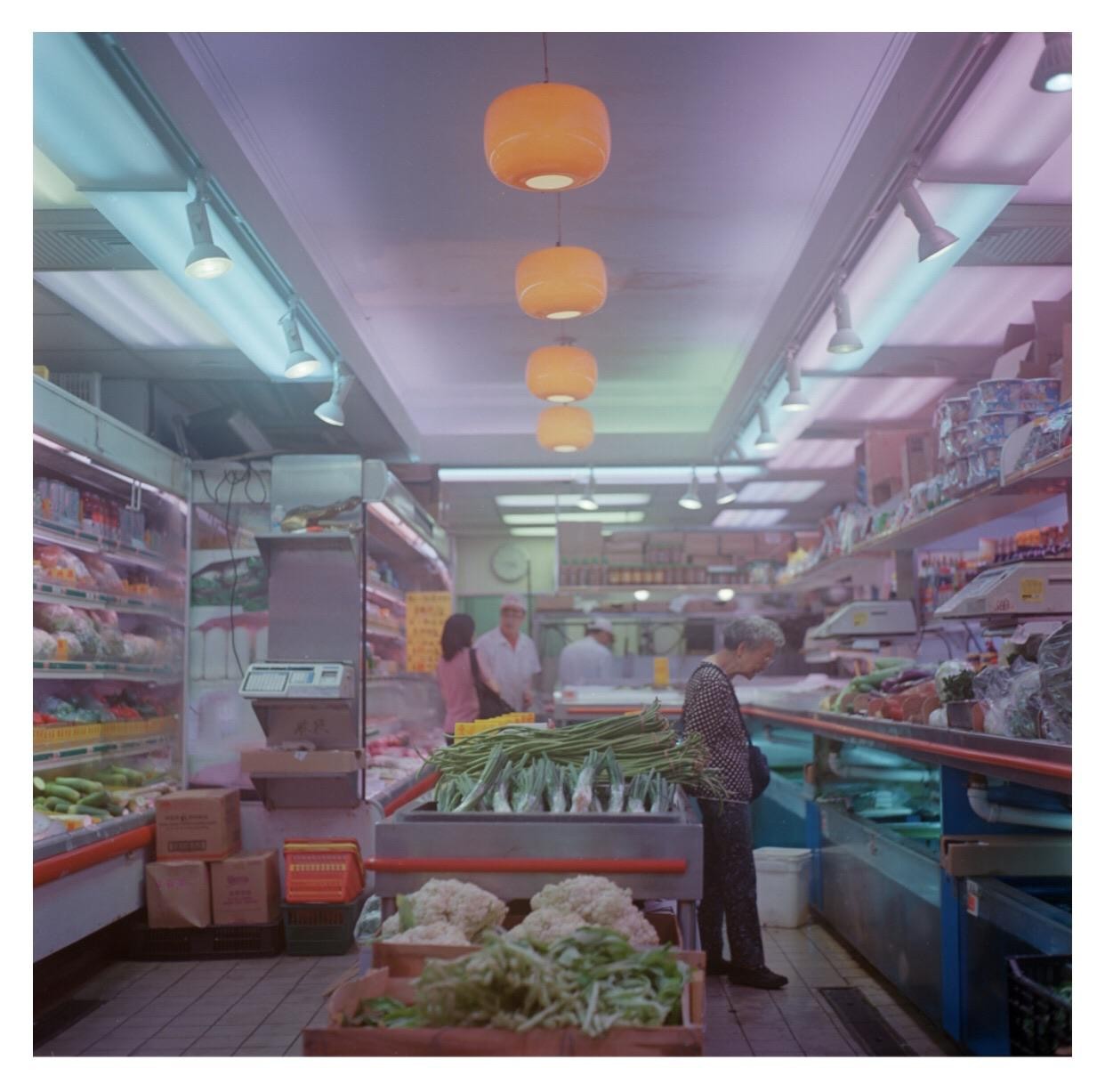 A woman shops for vegetables in a brightly lit grocery store with orange pendant lights.