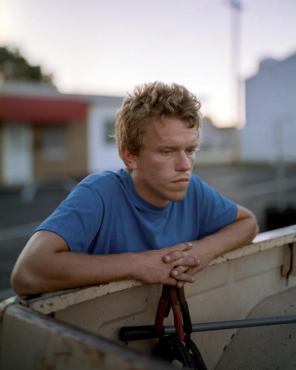 A man with red hair and a blue shirt leans on the back of a truck with jumper cables.