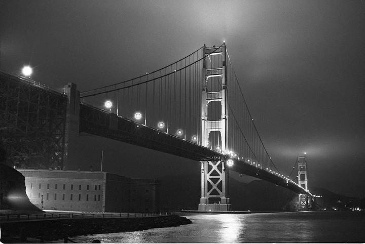 The Golden Gate Bridge is illuminated at night, with a building visible on the left shore and lights reflecting on the water.