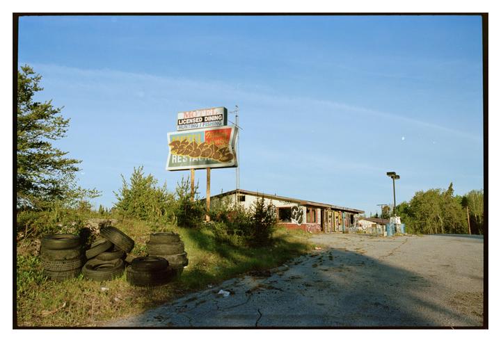 An abandoned motel with a large sign and piles of tires sits beside a cracked asphalt road.