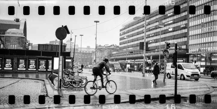 A cyclist rides past parked bicycles and a building with burger advertisements on a wet city street.