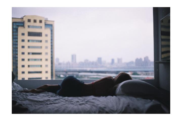 A woman lies on a bed looking out a window at a city skyline.