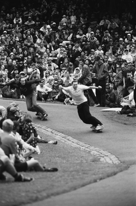 Two men on skateboards lean into a turn on a paved path while a large crowd watches from a grassy hill.